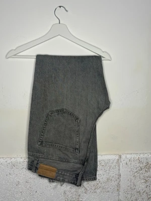 Grå raka jeans med bakficka - Säljer ett par grå jeans med rak passform och klassisk bakficka. Jeansen har en enkel design och en brun patch vid linningen. Perfekta för en avslappnad och stilren look.