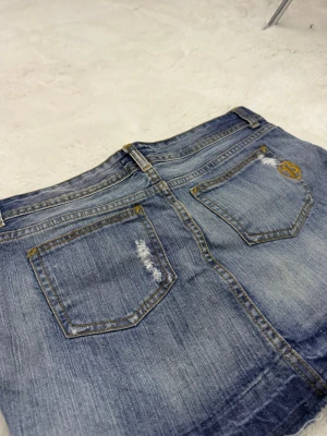 Blå jeanskjol med slitningar - Kort jeanskjol i blå denim med slitna detaljer och rå kant nertill. Kjolen har fem fickor, bälteshällor och stängs med knapp och dragkedja. Snygga kontrastsömmar och en liten broderad detalj på ena bakfickan. Säljer på grund av att den inte används längre❤️