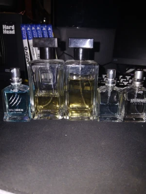 Fem herrparfymer från NG och Islande - Säljer ett paket med fem herrparfymer: två större flaskor från Islande (Gold Asset) med rektangulär form och svart kork, samt tre mindre flaskor från NG, varav en heter Wildmen (blå), en Homme (transparent) och en med blågrå ton. Alla flaskor är i glas.. men dom är danska mycket kvar men don är inte fulla som man ser på bilden