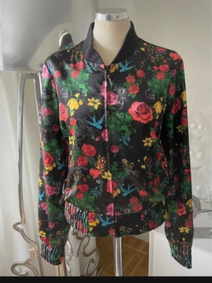 Svart bomberjacka med blommigt mönster - Snygg svart bomberjacka med färgglatt blommönster i rött, gult, grönt och blått. Jackan har dragkedja framtill, ribbade muddar och krage. Materialet ser glansigt ut och jackan har en avslappnad passform. Perfekt för dig som vill sticka ut med en unik stil.