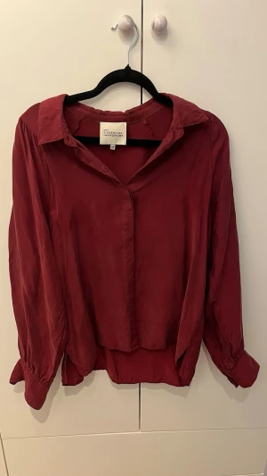 Vinröd blus från My Essential Wardrobe - Snygg vinröd blus i mjukt, glansigt material från My Essential Wardrobe. Klassisk krage, dold knappslå och långa ärmar med breda manschetter. Perfekt till jeans eller kjol för en trendig look.