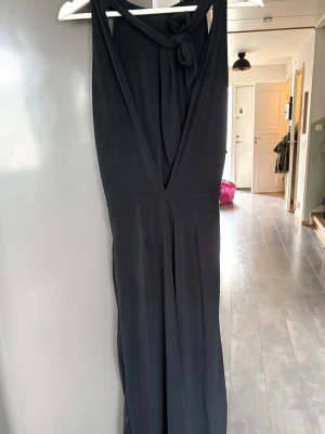 Svart ärmlös jumpsuit med djup rygg - Snygg svart jumpsuit med vida ben och  öppen rygg med knytband i nacken. Tillverkad i ett mjukt, fallande tyg som ger en elegant siluett. Perfekt för dig som vill ha en stilren och trendig look. 