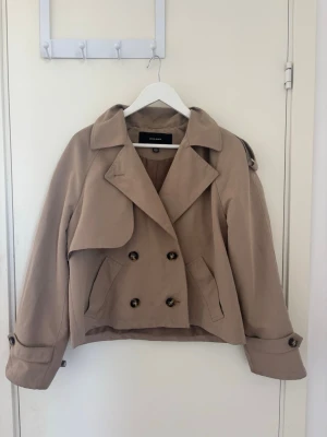 Kort beige trenchcoat från Veromoda  - Snygg kort trenchcoat från Veromoda i beige färg. Jackan har dubbelknäppning med stora knappar, klassisk krage och axelklaffar. Ärmarna har dekorativa spännen vid mudden och jackan har två fickor framtill. Perfekt för en trendig och stilren look.