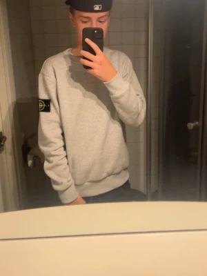 Grå sweatshirt från Stone Island - Säljer en stilren grå sweatshirt från Stone Island med den klassiska loggan på vänster ärm. Tröjan har rund halsringning, ribbade muddar och är tillverkad i mjukt bomullsmaterial. Perfekt för en avslappnad och trendig look.