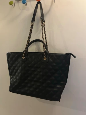 Svart handväska Don Donna - Snygg svart handväska från Don Donna i quiltat mönster. Väskan har kedjehandtag med gulddetaljer och dubbla bärhandtag. Tillverkad i konstläder och har en rymlig form som passar till mycket. Perfekt accessoar för dig som gillar stilrena detaljer. Väskan har  en slät sida och en mönstrad 