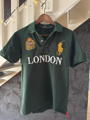 Grön Polo Ralph Lauren t-shirt London - Mörkgrön t-shirt från Polo Ralph Lauren med broderad gul logga och texten 'LONDON' framtill. Klassisk pikékrage och korta ärmar. T-shirten har även ett broderat emblem med brittisk flagga på bröstet. Tillverkad i bomull för skön känsla.