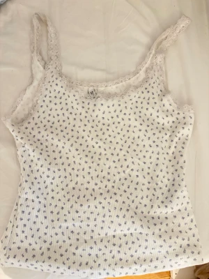 Vit blommig spetslinne - Söt vit ribbad linnetopp från DIVIDED med små blå blommor och spetskant vid urringningen. Perfekt för en somrig och fräsch look. Tunn och mjuk bomullskvalitet som känns skön mot huden.