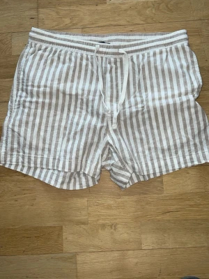 Randiga shorts i linne med snörning - Säljer ett par ljusbeige och vita randiga shorts i linne med elastisk midja och snörning framtill. Shortsen har avslappnad passform, sidofickor och en bakficka. 