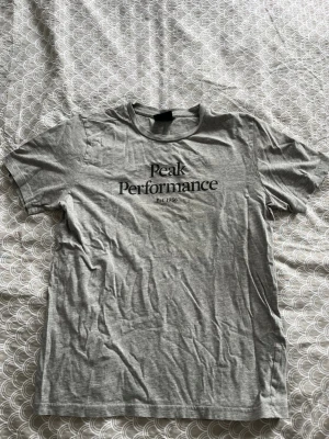 Grå Peak Performance t-shirt - Snygg grå t-shirt från Peak Performance med svart tryck på bröstet där det står 'Peak Performance Est. 1986'. Klassisk passform och rund halsringning. Perfekt basplagg till garderoben.