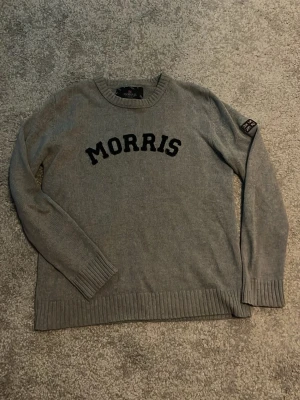 Grå stickad tröja från Morris - Snygg grå stickad tröja från Morris med svart text på bröstet och en patch med brittiska flaggan på ärmen. Tröjan har rund halsringning, långa ärmar och ribbade muddar. Perfekt för en avslappnad men stilren look.