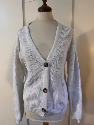 Vit stickad kofta med knappar - Mysig kofta med vanlig passform som går ganska långt ned. Köpt på en secondhandbutik men är aldrig använd av mig. Har en liten skada som syns på sista bilden. Passar även S. Skriv vid frågor!!