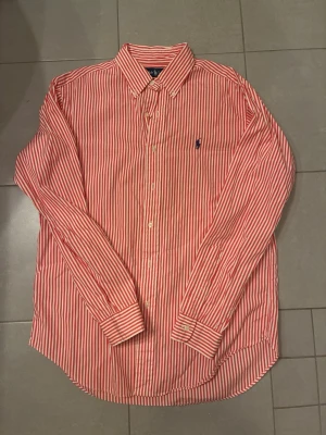 Rödvit randig skjorta från Polo Ralph Lauren - Klassisk långärmad skjorta från Polo Ralph Lauren med röda och vita vertikala ränder. Skjortan har button-down krage, knäppning framtill och den ikoniska blå broderade loggan på bröstet. Tillverkad i mjuk bomull för en bekväm känsla.