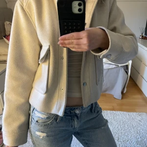 Beige vår jacka från Zara - Stilren beige jacka från Zara i kort modell med klassisk krage och stora fickor framtill. Jackan har knäppning med tryckknappar och är tillverkad i ett mjukt ull-liknande material. Perfekt att bära över en topp eller hoodie för en clean look.