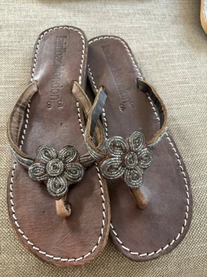 Sandaler med pärlor s38 - Helt NyA! Snygga bruna sandaler från Laidback London med dekorativ blomma av pärlor på remmen. Tillverkade i läder med vita kontrastsömmar runt sulan och tårem. Perfekta för en avslappnad och trendig sommarstil.