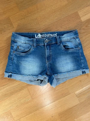 Blå jeansshorts från LAB Industries - Snygga blåa lågmidjade jeansshorts från LAB Industries med fransig kant och klassisk femficksdesign. Shortsen har normal passform, bälteshällor och knappgylf. Perfekta för varma dagar och en avslappnad stil.