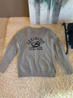 Grå Lexington sweatshirt med tryck - Snygg grå sweatshirt från Lexington med svart tryck framtill och klassiskt collegeinspirerat emblem. Tröjan har rund halsringning, ribbade muddar vid ärmslut och nederkant samt en relaxed passform. Perfekt för en casual och sportig stil.