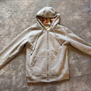 Grå hoodie från Burberry med dragkedja - Köpte i London för konfa present. Säljer för att den inte passar.                                                                                                        Snygg grå hoodie från Burberry med klassiskt rutigt foder i huvan. Tröjan har dragkedja framtill, snörning i huvan och två fickor. Perfekt för dig som vill ha en stilren och exklusiv look med en touch av Burberrys ikoniska mönster.