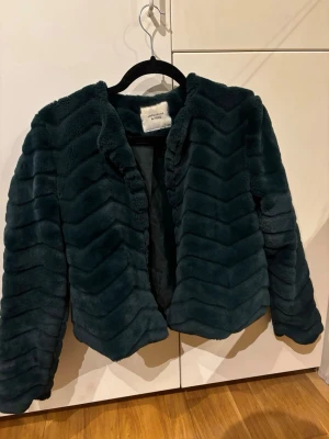 Mörkgrön pälsjacka från Jacqueline de Yong - Snygg mörkgrön pälsjacka från Jacqueline de Yong i mjuk fuskpäls. Jackan har ett vågigt mönster, öppen framtill och är midjekort med långa ärmar. Perfekt för att höja din vinterstil med en lyxig touch.
