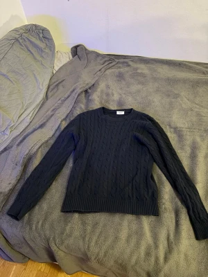 Mörkblå kabelstickad tröja Kappahl - Snygg mörkblå kabelstickad tröja från Kappahl i storlek 158/164. Tröjan har rund halsringning, långa ärmar och ribbade muddar vid ärmslut och nederkant. Perfekt för att hålla värmen under kyliga dagar.