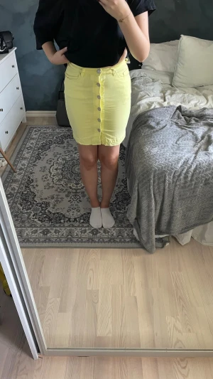Gul kjol med knappar framtill - Snygg gul kortkjol med knappar hela vägen framtill. Kjolen har en figurnära passform och är tillverkad i ett mjukt och stretchigt material. Perfekt till vår och sommar.