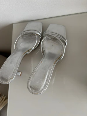 Silvriga pumps med smala remmar Zara - Säljer ett par silvriga pumps från Zara med smala remmar och fyrkantig tå. Skorna har låg klack och är tillverkade i ett glansigt material som ger en trendig look. Perfekta för dig som vill sticka ut med stilrena detaljer.