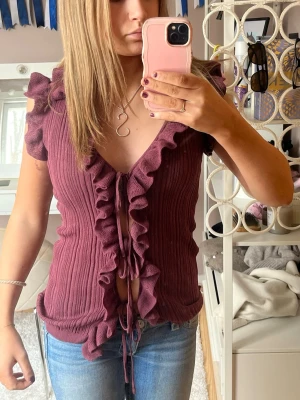 Vinröd ribbad topp med volang från Zara - Vinröd ribbad topp från Zara med volangkant längs hela framsidan och knytning framtill. Toppen är ärmlös med lätt rynkade axlar och har en croppad passform. Perfekt för dig som vill sticka ut med en trendig och söt look. Ingentligen storlek M men omsydd så sitter som en S!