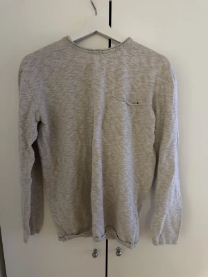 Ljusgrå långärmad tröja - En enkel och stilren ljusgrå långärmad tröja med rund halsringning och diskret bröstficka. Tröjan har en mjuk och lätt struktur, perfekt för lager på lager. Passar till både jeans och chinos för en avslappnad look. Den har dock ett pyttelitet hål över bröstfickan men inget märkbart (se bild), därav priset!