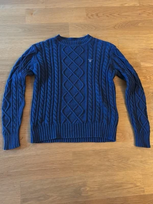 Blå kabelstickad tröja Gant  - Snygg blå kabelstickad tröja från Gant med rund halsringning och ribbade muddar. Tröjan har ett klassiskt flätmönster och en liten broderad logga på bröstet. Perfekt för dig som gillar stilrena och tidlösa plagg.                   (Frakten tillkommer)