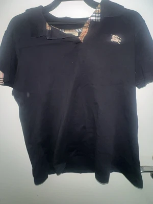 Svart Burberry t-shirt med rutiga detaljer - Snygg svart t-shirt från Burberry med klassiska rutiga detaljer vid kragen och ärmslut. T-shirten har korta ärmar, liten logga på bröstet och är gjord i mjuk bomull. Perfekt för dig som gillar stilrena plagg med exklusiv känsla. Står 3XL men sitter som S-xs