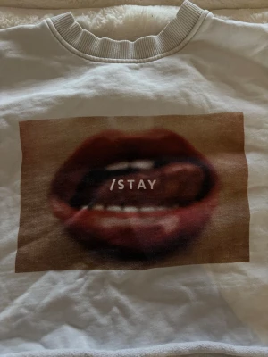 Vit sweatshirt med tryck /STAY - Säljer en vit sweatshirt med ett stort tryck av röda läppar och texten '/STAY' på bröstet. Tröjan har rund halsringning och ribbade muddar vid ärmslut och nederkant. Perfekt för dig som gillar statement-plagg och streetstyle. Köpt på carlings men har klippt av den längst ner.