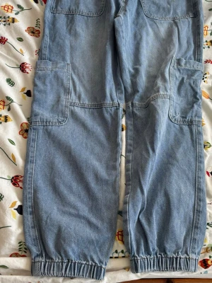 Baggy ljusblå cargojeans med resår - Baggy cargojeans i ljusblå denim med stora fickor på sidorna och resår vid bensluten. Perfekta för en avslappnad streetstil. Jeansen har en loose fit och är tillverkade i bomullsdenim.