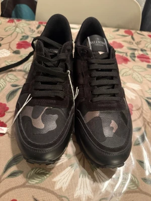 Svarta sneakers från Valentino - Snygga svarta sneakers från Valentino med coola kamouflagemönster på tån och metallicdetaljer på hälen. Skorna har svarta skosnören, robust sula och är gjorda i en mix av mocka och textil. Perfekta för dig som vill sticka ut med stilrena detaljer.