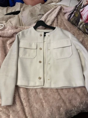 Vit kort cardiganjacka med fickor - Stilren vit cardiganjacka med rund halsringning och dekorativa guldfärgade knappar framtill. Jackan har två stora bröstfickor med lock och är tillverkad i ett mjukt, tjockare material. Passar perfekt till lager-på-lager och ger en clean look.