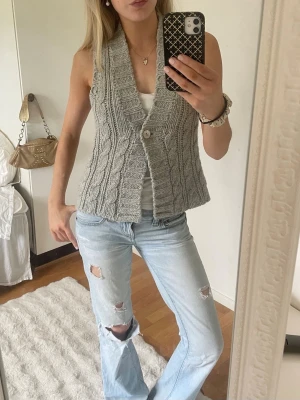 Grå stickad väst med kabelmönster - Säljer en grå stickad väst med kabelstickat mönster och bred krage. Västen har en knapp framtill och är ärmlös, perfekt att styla över en t-shirt eller skjorta. Materialet känns mjukt och mysigt, och modellen är något kortare. Storlek S