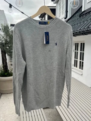 (Helt ny)Ljusgrå stickad tröja från Polo Ralph Lauren - (Moddelen är 187 och väger 77 kg )Snygg ljusgrå stickad tröja från Polo Ralph Lauren med rund halsringning och diskret blå logga broderad på bröstet. Tröjan har ribbade muddar vid ärmslut och nederkant, perfekt för lager på lager och enkel att matcha.