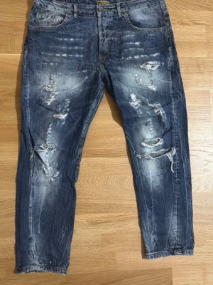 Blå slitna jeans med distressed detaljer - Säljer ett par blå jeans med coola slitningar och distressed detaljer framtill. Jeansen har normal passform, klassisk femficksdesign och är tillverkade i bomull. Perfekta för en avslappnad och trendig look.
