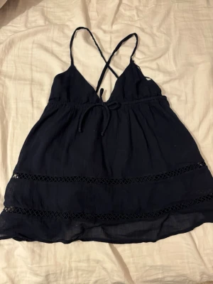 Mörkblå linnetopp med spetsdetaljer - Snygg mörkblå linnetopp med tunna axelband som korsas i ryggen och spets detaljer nertill. Säljs då den blivit för liten.