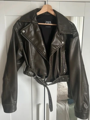  skinnjacka från Zara - Snygg svart skinnjacka från Zara med klassisk bikerstil. Jackan har breda slag, silverfärgade dragkedjor och detaljer samt ett bälte i midjan. Croppad modell som ger en cool och trendig look. Perfekt för dig som vill ha en edgy vibe, kommer ej till användning, ganska liten i storleken💕