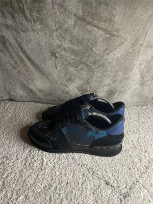 Valentino rockrunners  - Snygga sneakers från Valentino Garavani i svart och blått med kamouflagemönster. Skorna har lågt skaft, svarta skosnören och detaljer i mocka och textil. Perfekta för dig som vill ha en unik och trendig look.SULAN HAR LÅSNAT  FRÅN SKORNA LITE