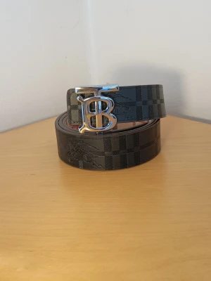 Svart rutigt Burberry bälte - Snyggt svart bälte från Burberry med klassiskt rutigt mönster och en stor silvrig B-spänne framtill. Bältet är gjort i skinn och har en exklusiv känsla som lyfter vilken outfit som helst.