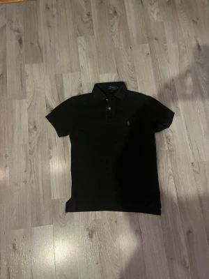 Svart pikétröja från Polo Ralph Lauren - Klassisk svart pikétröja från Polo Ralph Lauren med korta ärmar och krage. Tröjan har två knappar framtill och den ikoniska broderade loggan på bröstet. Tillverkad i mjuk bomull för en bekväm känsla.