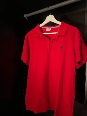 Röd pikétröja från Polo Ralph Lauren - Klassisk röd pikétröja från Polo Ralph Lauren med korta ärmar och krage. Tröjan har knappar vid halsen och den ikoniska broderade loggan på bröstet. Perfekt till jeans eller shorts för en avslappnad men snygg look.