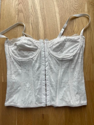 Vit spetskorsett med snörning - Snygg vit korsett med spetsdetaljer och justerbara axelband. Framsidan har hyskknäppning och baksidan har snörning.