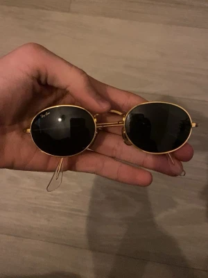 Ray-Ban ovala solglasögon guld - Snygga ovala solglasögon från Ray-Ban med tunna guldfärgade metallbågar och mörka linser. Klassisk och tidlös design som passar till många stilar. Medföljer ett svart fodral med Ray-Ban-logga i guld.