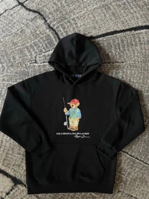 Polo Ralph Lauren hoodie - Svart hoodie från Ralph Lauren med Polo Bear-tryck framtill där björnen har golfkläder och röd keps. Nypris 2200kr | mitt pris 749kr | aldrig tvättad eller så. Använd 2 gånger. Helt ny och perfekt för dig som vill ha en snygg hoodie!! Skriv vid fundering eller fler bilder!
