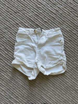 Vita högmidjade jeansshorts - Säljer ett par vita högmidjade jeansshorts med uppvikta benslut. Shortsen har klassisk femficksdesign, knapp och dragkedja framtill samt bälteshällor. Perfekta för varma dagar och enkel att matcha med allt.