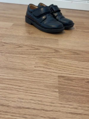 Svarta loafers med kardborreband - Stilrena svarta loafers i blankt läder med breda kardborreband över vristen. Skorna har rundad tå och låg sula, vilket ger en klassisk look. Insidan är ljusbrun och materialet ser ut att vara äkta läder. Perfekta för dig som gillar enkel och tidlös stil.