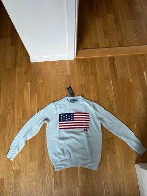 Ljusblå stickad tröja Polo Ralph Lauren - Ljusblå stickad tröja från Polo Ralph Lauren med amerikansk flagga broderad på bröstet och RL-detalj. Tröjan har rund halsringning, ribbade muddar och är tillverkad i bomull för en skön känsla.