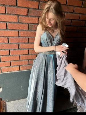 Silvrig balklänning  - Säljer en magisk långklänning med glittrigt tyg som fångar ljuset superfint. Klänningen har en vid, svepande kjol och är ärmlös, perfekt för bal eller student. Materialet känns lyxigt och skimrande, och färgen drar åt silverblått. Ingen synlig logga.