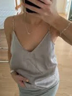 Ljusgrå silkeslinne med kedjeband - Säljer ett ljusgrått linne i silkesliknande material med tunna kedjeband och djup v-ringning. Snyggt och stilrent plagg som passar perfekt till jeans eller kjol. Linnet har en avslappnad passform och känns lyxigt mot huden. Knappt använd (lånad första bild)💕